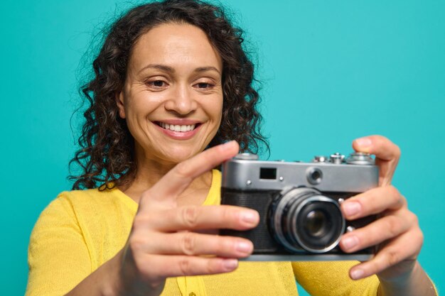 Cheerful African American woman photographer in bright yellow clothes smiling toothy smile while holding an old retro style vintage camera and photographing, taking pictures against blue background