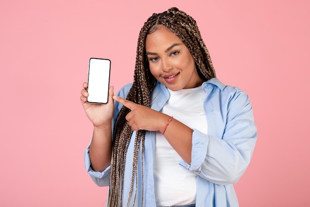 Веселая афроамериканка показывает мобильный телефон на розовом фоне