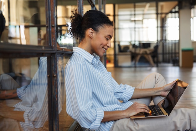 cheerful african american woman smiling and typing on laptop while sitting on floor in modern office