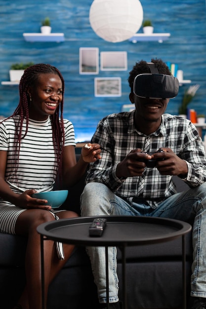 Cheerful african american woman watching boyfriend playing VR game simulation in living room while eating a bowl of snacks. Content young adults at home enjoying virtual reality cyberspace using high