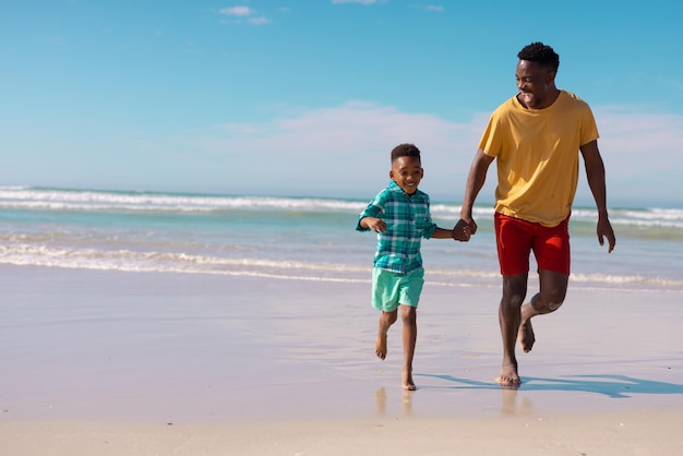 Cheerful african american young man and son holding hands while running at beach against blue sky