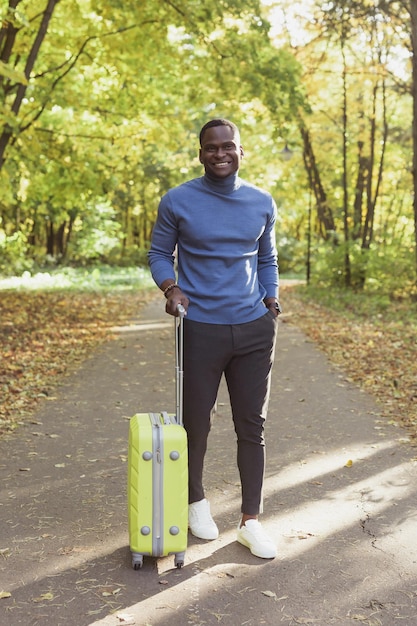 Cheerful african american young man in stylish clothes with suitcase walks in an spring park on warm