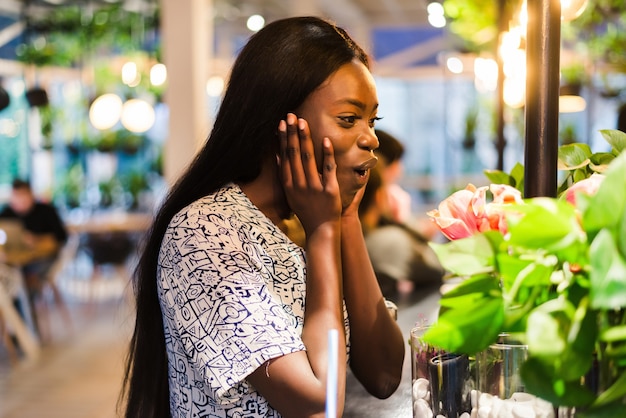 カフェで夏のドレスを着た陽気なアフリカ系アメリカ人の若い女性は、花瓶の白い花を嗅ぎます。