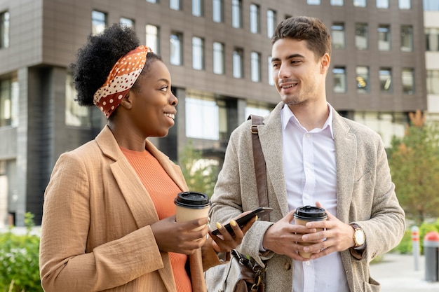 Веселая африканская бизнесвумен с напитком смотрит на коллегу во время прогулки в городской среде и обсуждает хорошие новости