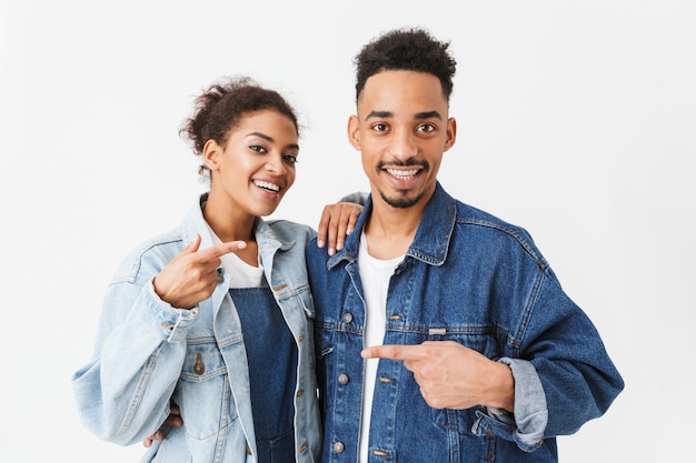 Cheerful african couple in denim shirts pointing to each other  over grey wall