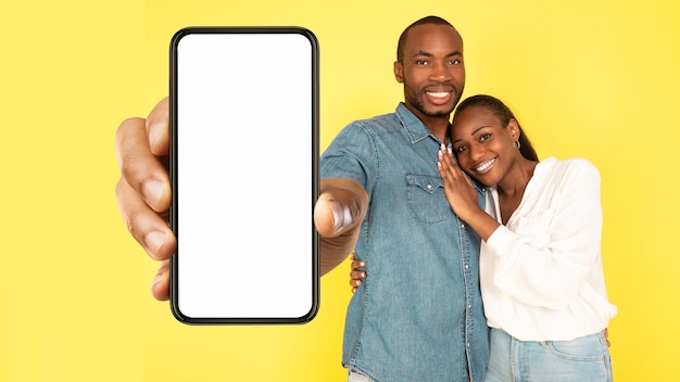 Cheerful African Couple Showing Large Cellphone Screen On Yellow Background