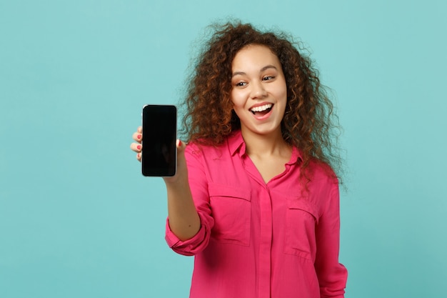 Cheerful african girl in casual clothes hold mobile phone with blank empty screen isolated on blue turquoise wall background in studio. People sincere emotions, lifestyle concept. Mock up copy space.