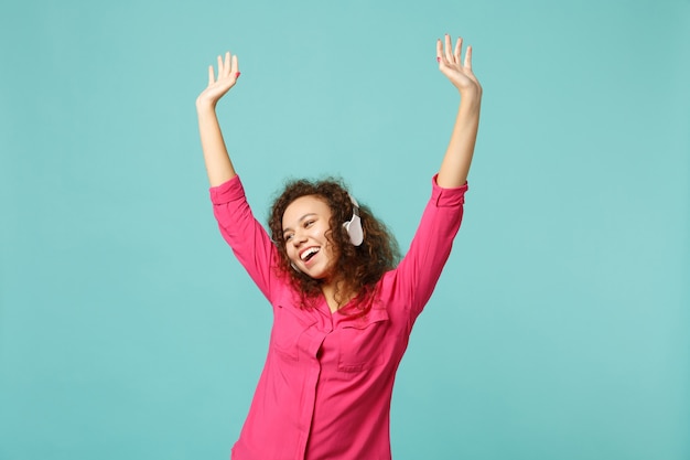 Cheerful african girl in pink casual clothes listening music with headphones and dancing isolated on blue turquoise wall background. People sincere emotions, lifestyle concept. Mock up copy space.