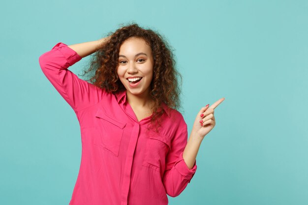 Ragazza africana allegra in abiti casual rosa che punta il dito indice da parte mettendo la mano sulla testa isolata sul fondo della parete turchese blu. concetto di stile di vita di emozioni sincere della gente. mock up copia spazio.