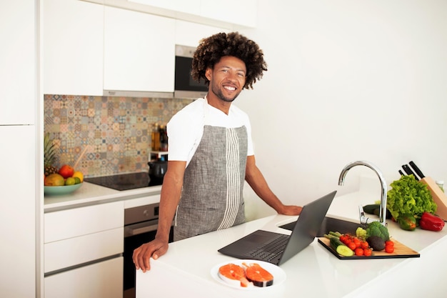 写真 室内で調理するデジタルレシピをナビゲートするラップトップで陽気なアフリカ人男性
