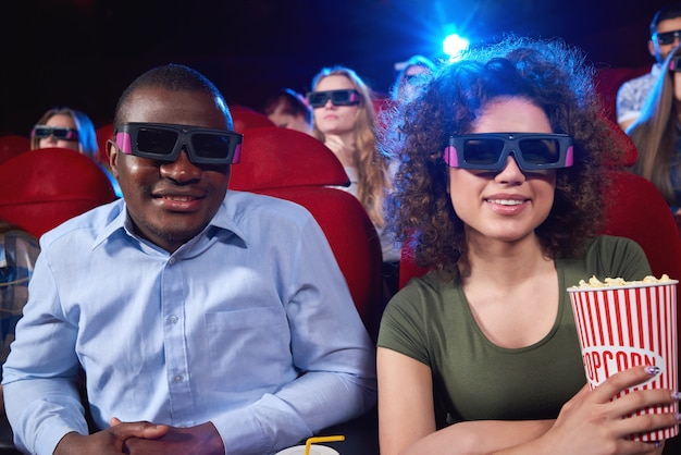 Cheerful African man and his girlfriend wearing 3D glasses holding popcorn smiling while enjoying a movie together couples dating romance friends friendship leisure entertainment.