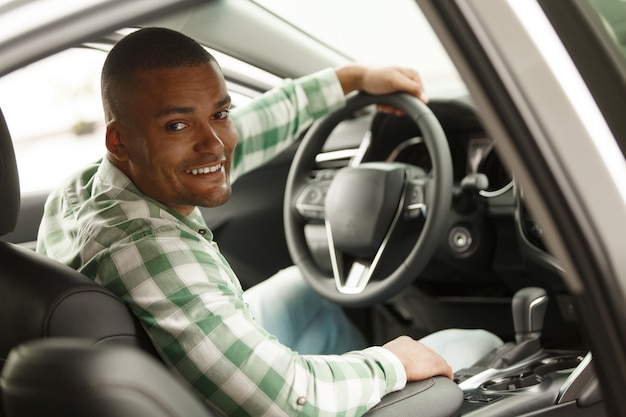 Cheerful African man smiling to the camera, sitting in his new car at the dealership. 