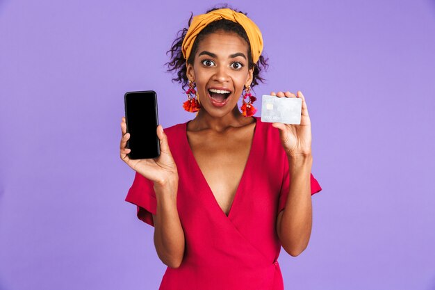 Cheerful african woman in dress holding credit card and showing blank smartphone screen