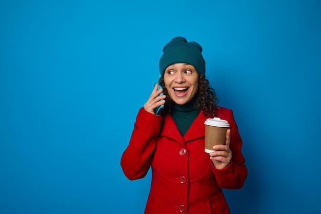 Cheerful African woman dressed in bright red coat and green warm woolen hat talks on mobile phone and holds a disposable cardboard paper mug with hot drink. Blue background copy space