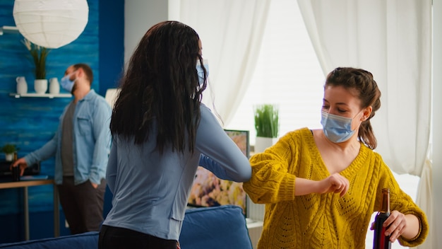 Cheerful african woman and her friend touching elbow keeping social distancing while greating each other wearing face mask, to prevent coronavirus spread in the course of global pandemic in living roo