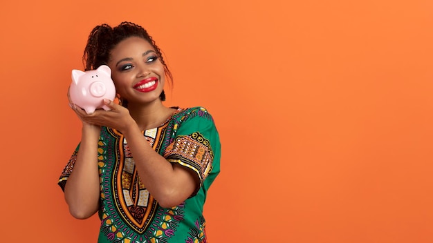 Cheerful african woman holding piggy bank next to ear