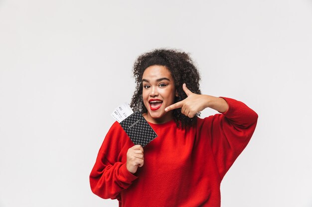 Cheerful african woman in red sweater holding passport with tickets and pointing on it while looking directly over grey background