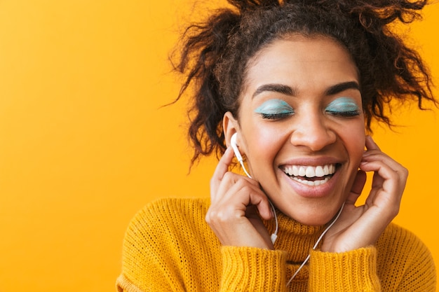 Cheerful african woman wearing sweater standing isolated, listening to music with earphones