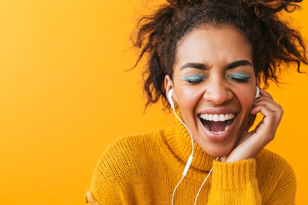 Cheerful african woman wearing sweater standing isolated, listening to music with earphones