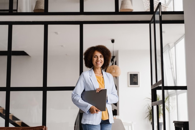 Cheerful african young woman programmer is standing with laptop\
and smiling candid african american girl with laptop standing in\
office smiles and rejoices at successful work in it company
