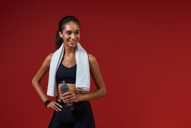 Cheerful afro american woman in sports clothing with towel on her shoulders holding bottle of