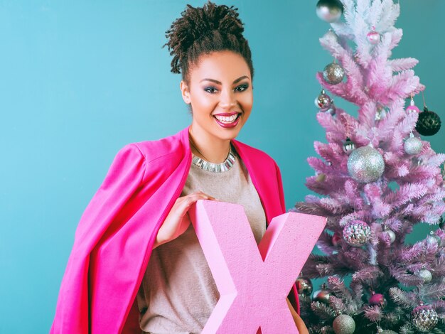 cheerful afro american woman with pink letter X in her hands on the christmas tree background