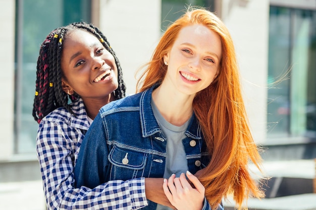 Donne lesbiche afro ed europee allegre che ridono e che guardano la macchina fotografica alla città del centro di estate