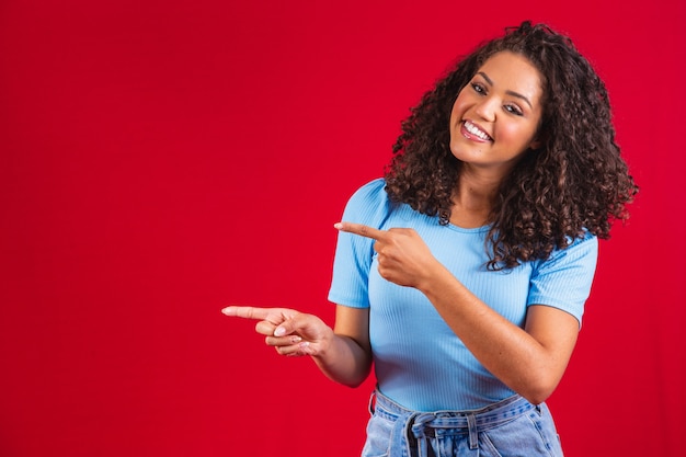 Cheerful afro woman points away on copy space, discusses\
amazing promo, gives way or direction, wears yellow warm sweater,\
has pleasant smile, feels optimistic, isolated over red\
background.