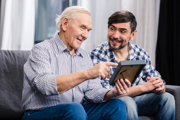 cheerful aged father and his son sitting on the sofa while holding a photo frame