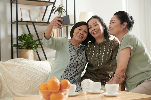Cheerful aged woman taking selfie with her best friends when they are having small party at home