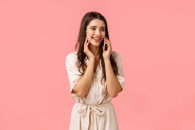 Cheerful alluring and carefree happy woman in dress, laughing talking on phone, calling best friend and discuss romantic date, holding smartphone near ear, look sideways, pink wall