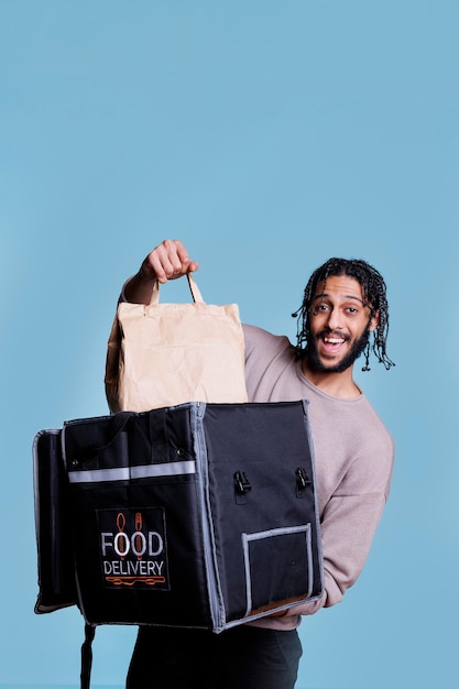 Веселый арабский доставщик берет посылку с едой из теплового портрета рюкзака. Счастливый курьер службы доставки ресторана раздает бумажный пакет с едой на вынос и смотрит в камеру