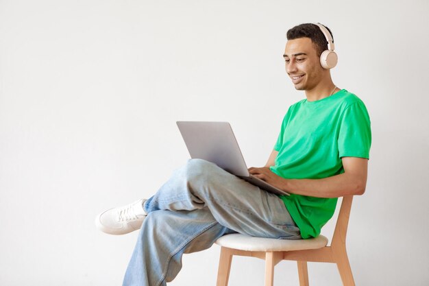 Cheerful arab man working on laptop and wearing headphones
sitting on chair over light studio wall copy space