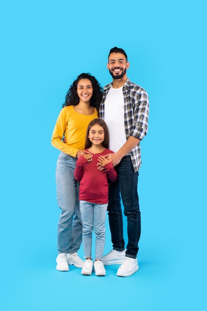 Cheerful arabic family of three posing embracing blue background vertical