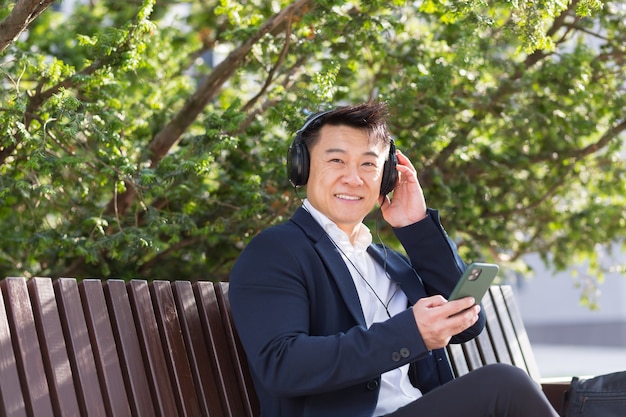 쾌활한 아시아 사업가는 시내 공원의 벤치에 앉아 손에 전화를 들고 헤드폰으로 음악을 듣고 있습니다. 정장을 입은 남성은 야외 활동을 즐깁니다. 긴장을 풀고 휴식을 취하십시오. 생활 양식