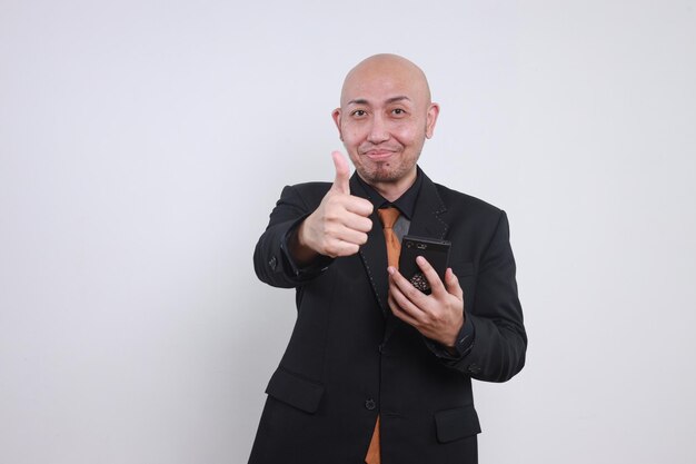 Photo cheerful asian businessman in formal suits showing tumbs up at camera while holding smartphone