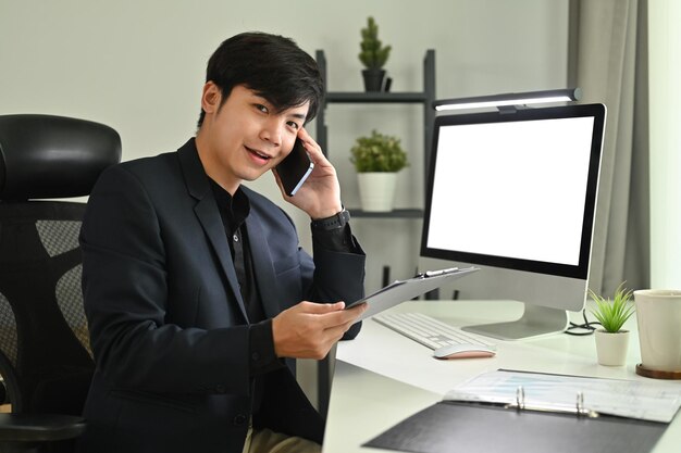 空のコンピュータ画面の前で座って携帯電話で話している陽気なアジア人ビジネスマン