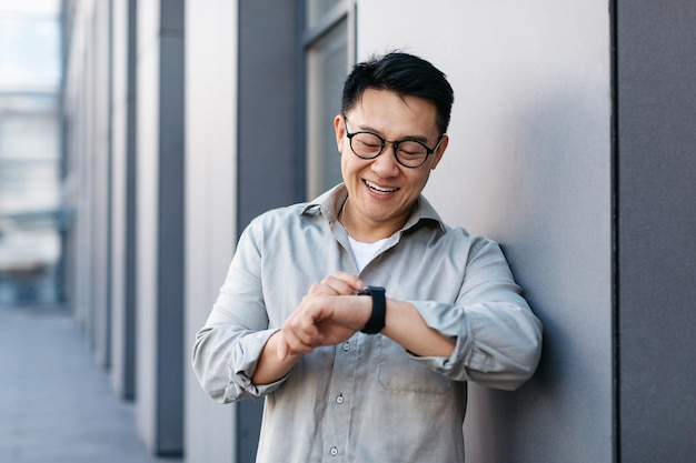 Cheerful asian businessman wearing smart watch and looking at it checking time using modern gadget outdoors