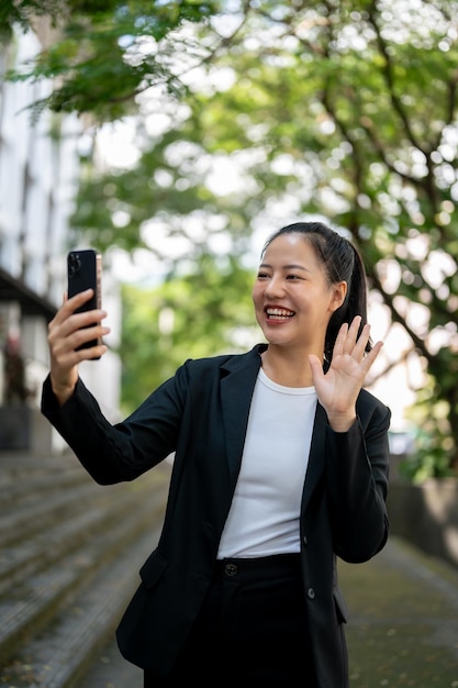 A cheerful Asian businesswoman is waving her hand while having a video call on her phone outdoors