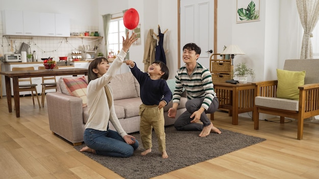 cheerful asian dad, mom and son having fun playing hit the ball game in the living room at home. they try to keep the balloon in air