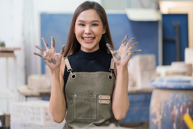 Cheerful asian female ceramist show hand dirty while sculp\
molding clay vase in home studio factory with happiness proudly\
smiling look at cameraasia female wear apron hand gesture smile in\
factory