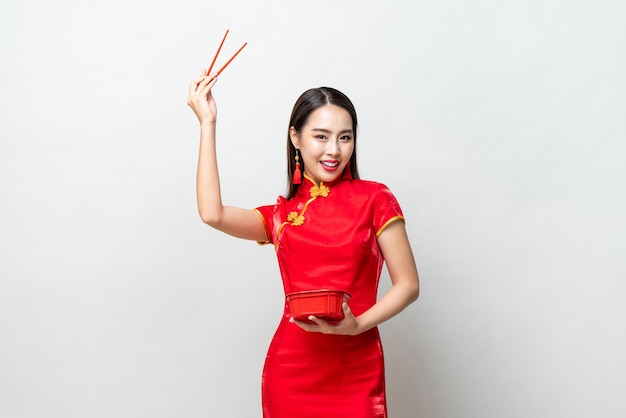 전통적인 빨간 드레스를 입은 쾌활한 아시아 여성이 젓가락으로 팔을 들고 회색 배경에 맞서기 위해 접시를 먹기 전에 미소로 카메라를 바라보고 있습니다.