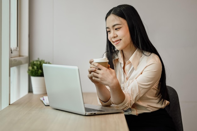 カフェでコーヒーを飲みラップトップでメールを読んでいる陽気なアジア人女性オフィスワーカー