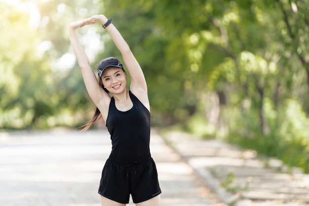 A cheerful Asian female runner in sports outfits doing stretching before jogging exercise outdoor in the city natural park under evening sunset