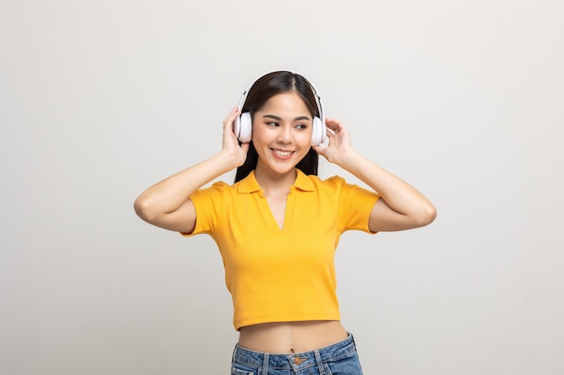 쾌활한 아시아 여성 십대는 격리된 흰색 헤드폰으로 음악을 듣습니다. 노란색 셔츠를 입은 아름다운 젊은 여성이 방에서 음악과 함께 즐거운 시간을 보내는 무선 헤드폰을 만집니다.