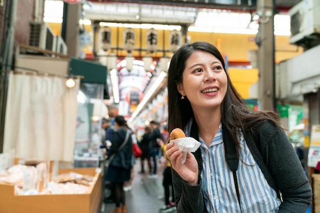 Photo cheerful asian girl backpacker holding eating a donut is having fun visiting kuromon ichiba market in osaka japan. she looks into space with smile on face.