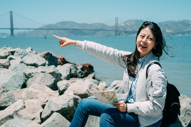 Il viaggiatore con zaino e sacco a pelo asiatico allegro della ragazza che tiene una mappa sta sorridendo alla macchina fotografica mentre indica a distanza dal mare con il ponte di golden gate sullo sfondo nella soleggiata california.