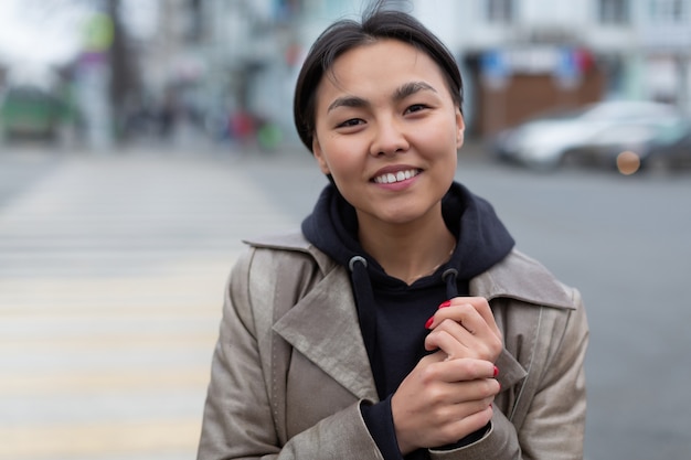 Веселая азиатская девушка в бежевом пальто и черном пиджаке с ярким маникюром стоит среди города улыбается