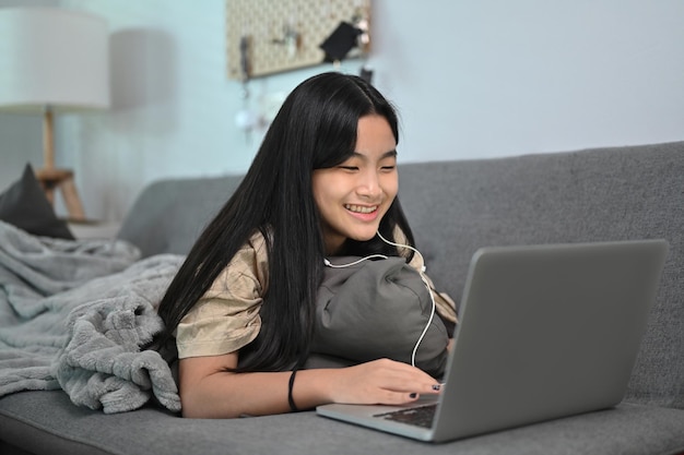 Cheerful asian girl lying on couch and surfing internet wit laptop computer.