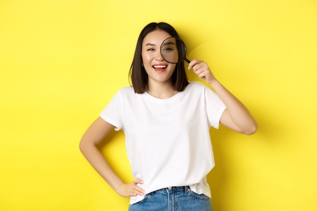 Photo cheerful asian girl searching for you, looking through magnifying glass and smiling, found something interesting, standing over yellow background.